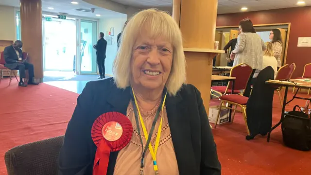 A woman with short, light-coloured hair wears a suit jacket and a peach-coloured top. She has a large red rosette on her jacket.