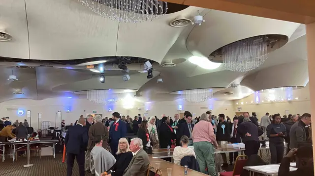 People sitting around in a hall at an election count under a chandelier
