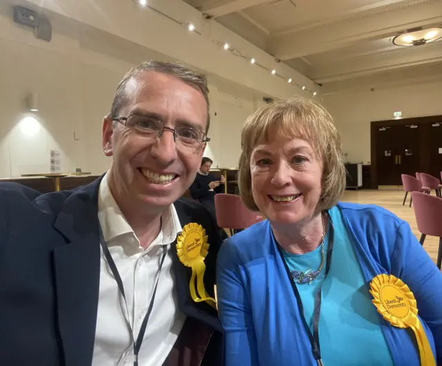 A man and woman posing together with smiles inside a large hall. Bother wearing yellow lib dem rosettes.