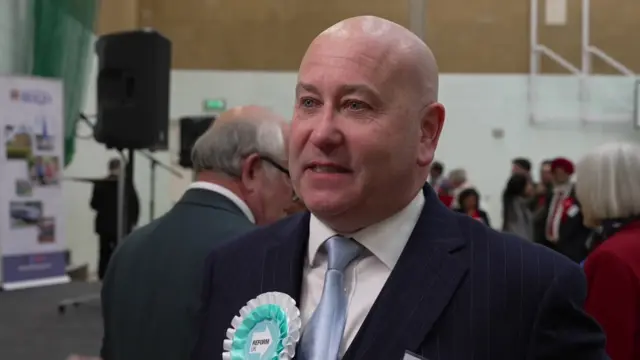 A man in a suit and tie with a Reform UK rosette being interviewed at an election count.