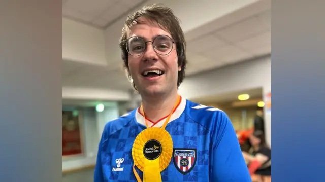 Paul Edgeworth is smiling with his mouth open and looking right at the camera. He has short brown hair and round glasses. He is wearing a blue football shirt with a yellow rosette around his neck.