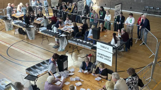 People at an election count in a leisure centre. Many sit at desks counting votes and others stand behind metal railings.
