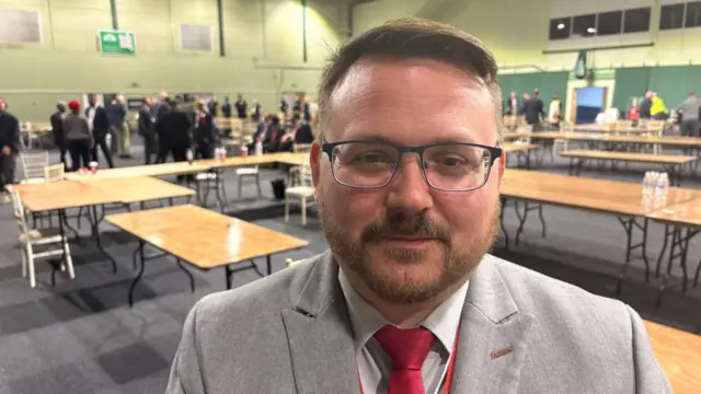 A man wearing a light grey suit, red tie and glasses looks at the camera while at an election count