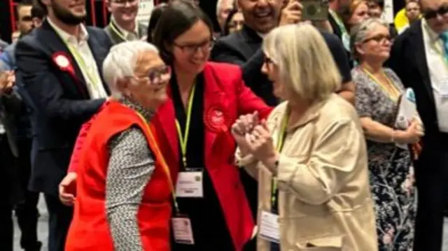 Three women hug as they celebrate a win. They are wearing lanyards and others stand behind them. One is in a red gilet, another a red jacket and the third a beige jacket.