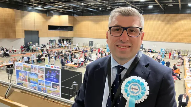 A man in a suit wearing a “Reform UK” rosette stands overlooking a busy hall where election staff sort ballot papers at long tables below.