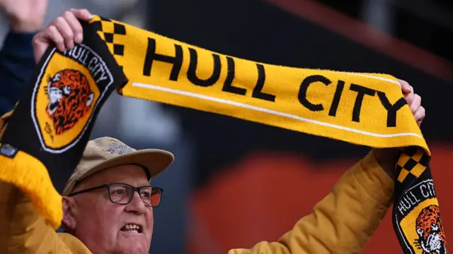 A Hull City fan wearing a baseball cap and holding aloft a club scarf with the team name on it