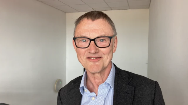 A man with short hair smiles for the camera wearing black framed glasses and a grey suit jacket over a blue and blue and white pinstriped shirt