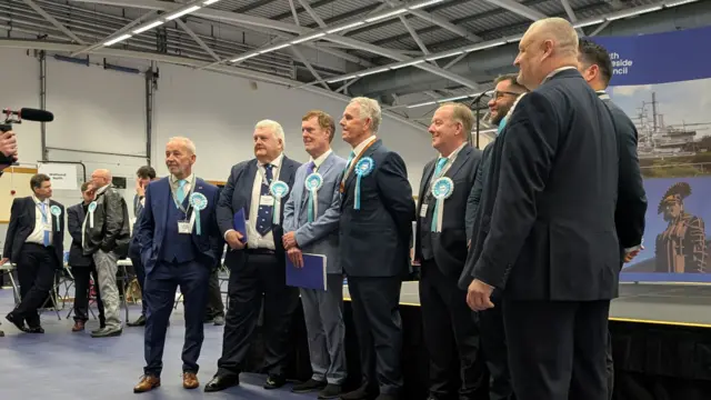 Eight men stand in a line in front of the stage and smile for the photographer in front of them. They are all wearing turquoise rosettes for Reform.