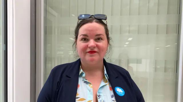 Heather Williams stares at the camera. She wears a navy jacket with a Conservative sticker on it. She wears a flowery dress and has glasses on her head