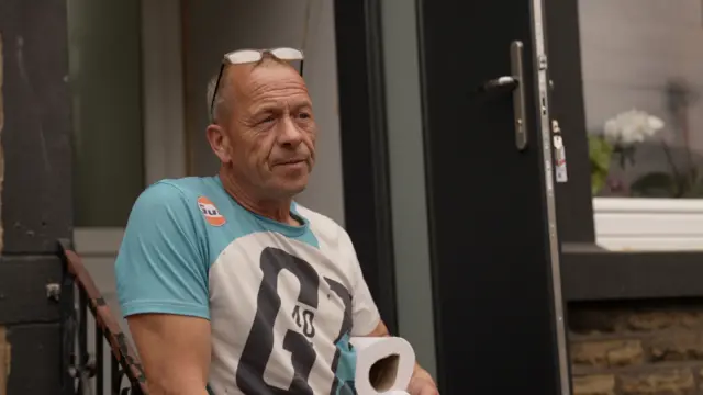 Lee sits in a blue t-shirt in front of a door.