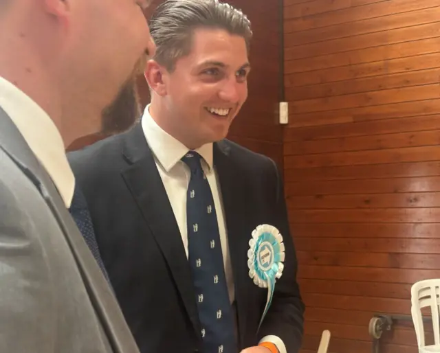 A man in a navy suit and tie with white shirt and sky blue Reform UK rosette smiling. He has brown hair combed back