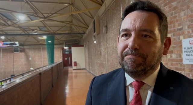 David Baines has dark hair and a beard. He wears a navy suit jacket with a red tie and white shirt