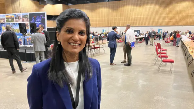 A woman is standing and facing the camera. She is wearing a dark blue blazer over a light-coloured top, along with a visible lanyard and credential badge hanging from the neck. The badge includes partially readable text, with “Candidate” and “Counting Agent” visible, suggesting a role connected to an organised process or formal event. The individual is positioned slightly off-centre and occupies a prominent portion of the frame.