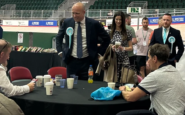 Dan Thomas in dark suit with cycling track in background standing at table talking to people seated