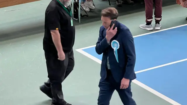 Andrew Husband is walking through the sports hall with a phone held to his ear. He is wearing a turquoise rosette for Reform UK and a blue suit.