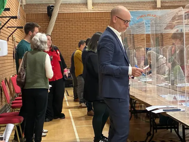 Stephen Kinnock in navy suit and glasses standing at table holding pen and paper