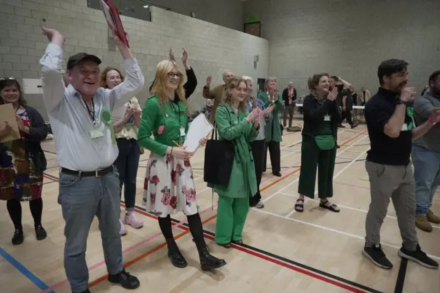 A group of men and woman clap and smile in a sports hall wearing green items of clothing to symbolise their affiliation to the Green Pary