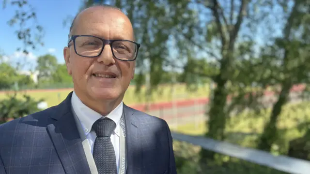 A man smiles at the camera standing outside on a sunny day. He is largely bald and wears a navy suit with a white shirt and tie as well as glasses.