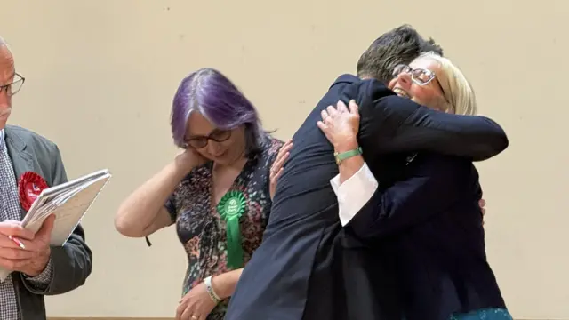 A woman and a man hug during an election vote count. The woman has blonde hair, glasses and wears a blue top with a black candidate.