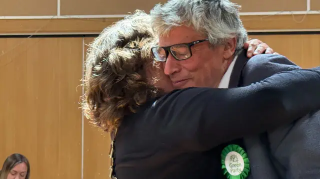 A man and woman embrace in a hug. The man has grey hair and wears a navy suit with a green rosette attached. The woman's face can't be seen but she wears a black top and has short brown, curly hair.