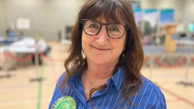 A woman with shoulder length brunette hair and black framed glasses smiles for the camera wearing a blue shirt and a green political rosette, with an empty polling station seen in the background in soft focus