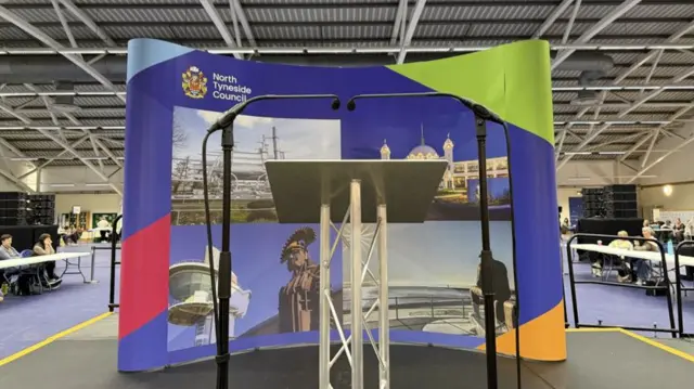 The small black stage has two microphones and a podium on the front. There is a large North Tyneside Council banner behind with various pictures of the area including the white domes of Spanish City.