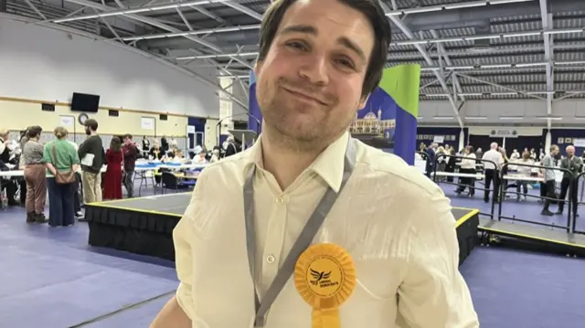 Alexander Martin is shrugging with a tight smile on his face. He is wearing a yellow shirt and rosette. Behind him, people are standing around the sports hall which has a stage in the middle.