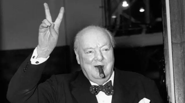 A black and white image of Winston churchill smoking a cigar and giving the v sign of victory