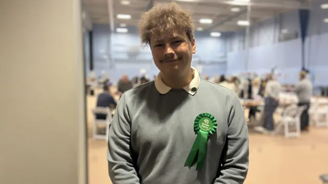 Connor Edward Shaw stood in a hall, wearing a green rosette on his chest.