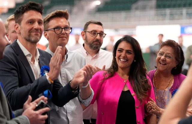 Natasha Asghar is wearing a pink top and is surrounded by people clapping her
