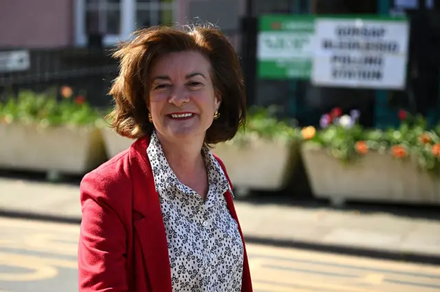 Eluned Morgan in red blazer and white patterned shirt walking in street smiling at camera