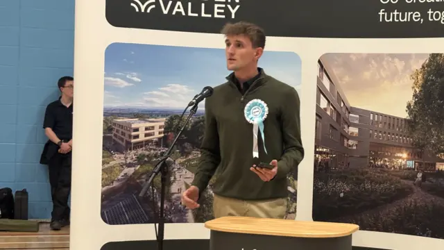 A man wearing a green jumper and a blue Reform rosette, standing behind a podium and speaking into a microphone.