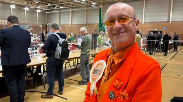 A man smiles at the camera in an orange suit