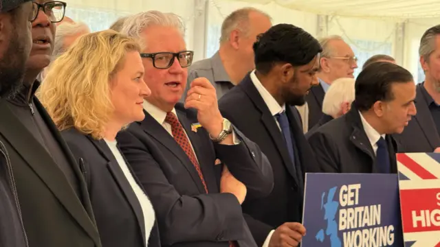 A man and a woman stand together and talk. They are wearing suits and are standing near signs which say Get Britain Working