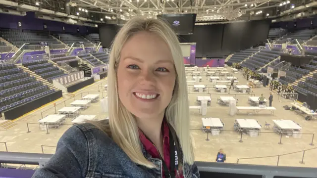 A blonde woman smiling in front of an empty room with lots of tables laid out.