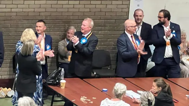People are standing behind a table clapping. Most of them are men and they are wearing blue rosettes.