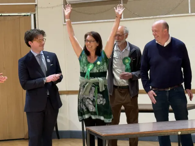 A woman celebrates with her hands in the area. She has short dark hair, glasses and wears a green top. Three men surround her and smile.