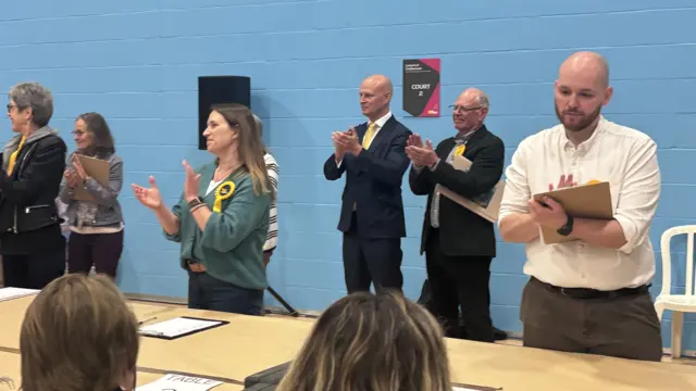 A row of councillors standing behind a wooden table in a large hall, clapping and cheering as results are read out.