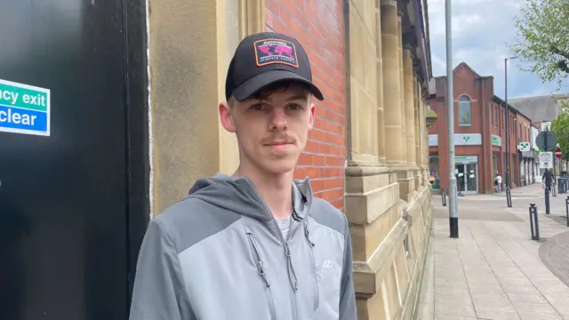 Luke is pictured outside a red brick building on a pavement, wearing a grey hooded jacket and a black cap