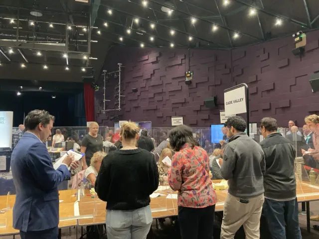 People gathered at a local election count inside a large hall.