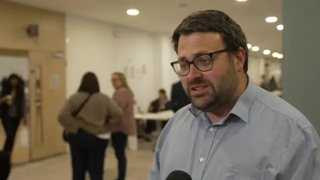 A middle aged man in a blue shirt is being interviewed at an election count.