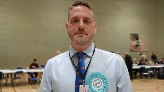 A man with blond hair and beard wearing a blue shirt and a blue tie. The tie has union flags on it. He is wearing a blue Reform UK rosette