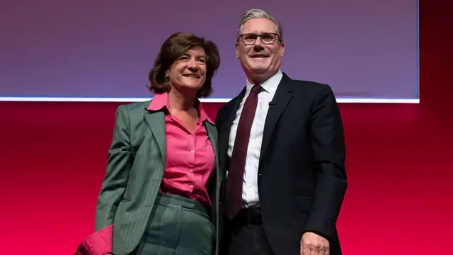Eluned Morgan (L) in a green suit and hot pink shirt smiles as she poses with Keir Starmer (R) at the Welsh Labour Party Conference. They're standing on a stage, a red wall with a clear screen behind them