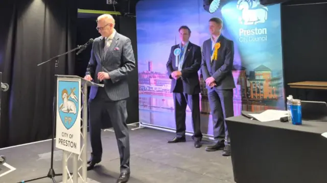 A man in a suit stands before a lecturn with the crest of Preston City Council on it