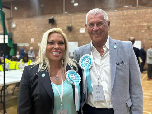 Emma Beck and Tony Keogh smile for the cameras after being elected to St Helens Council. They are both wearing turquoise and white rosettes in Reform UK's colours