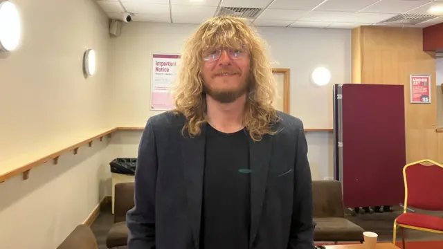 A man with long, shaggy blonde hair wearing a black suit and glasses smiles at the camera.