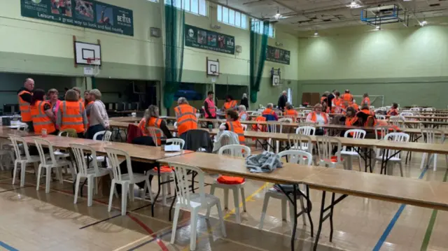 Lots of people in high-vis jackets inside a sports hall among tables and chairs