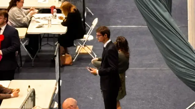 A man in a suit stands looking at people counting election ballots