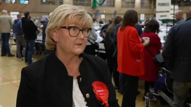 Tracey Dixon is speaking to an interviewer off camera. She has short blonde hair and glasses. She is wearing a dark blazer and a red rosette which reads: "Labour."