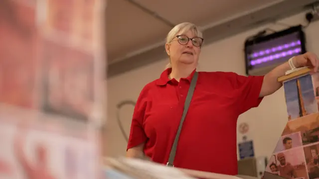 Adele stands in a red shirt behind a counter in a shop